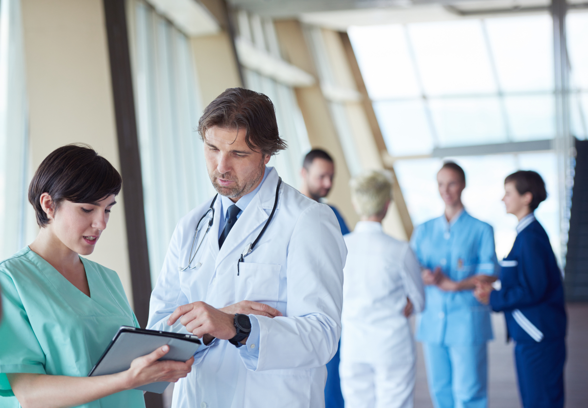 A doctor and a nurse going over detials on a tablet with people standing behind them in the foreground
